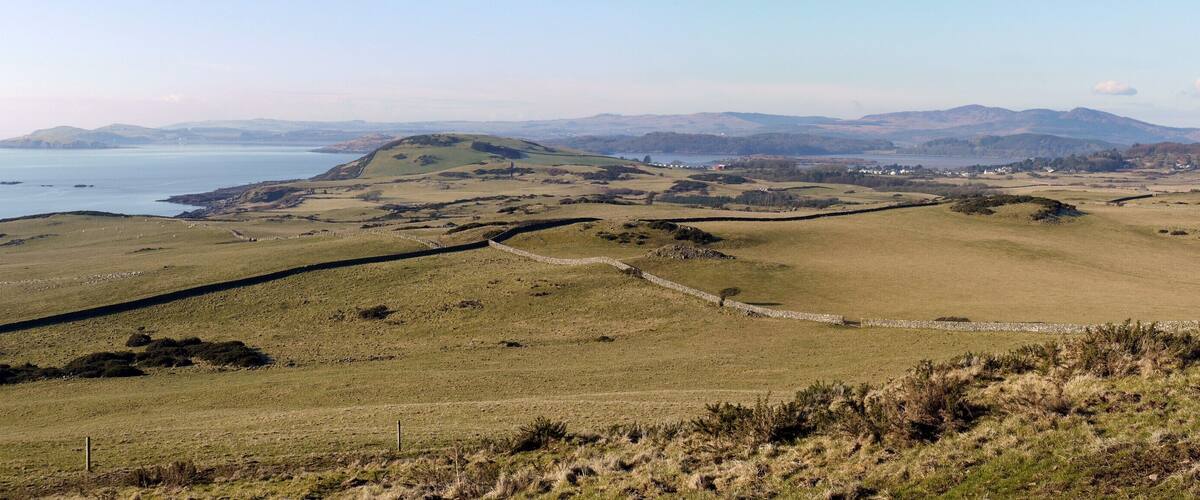 A coastal landscape with rolling hills, a stone wall, and a distant view of the sea and mountains.