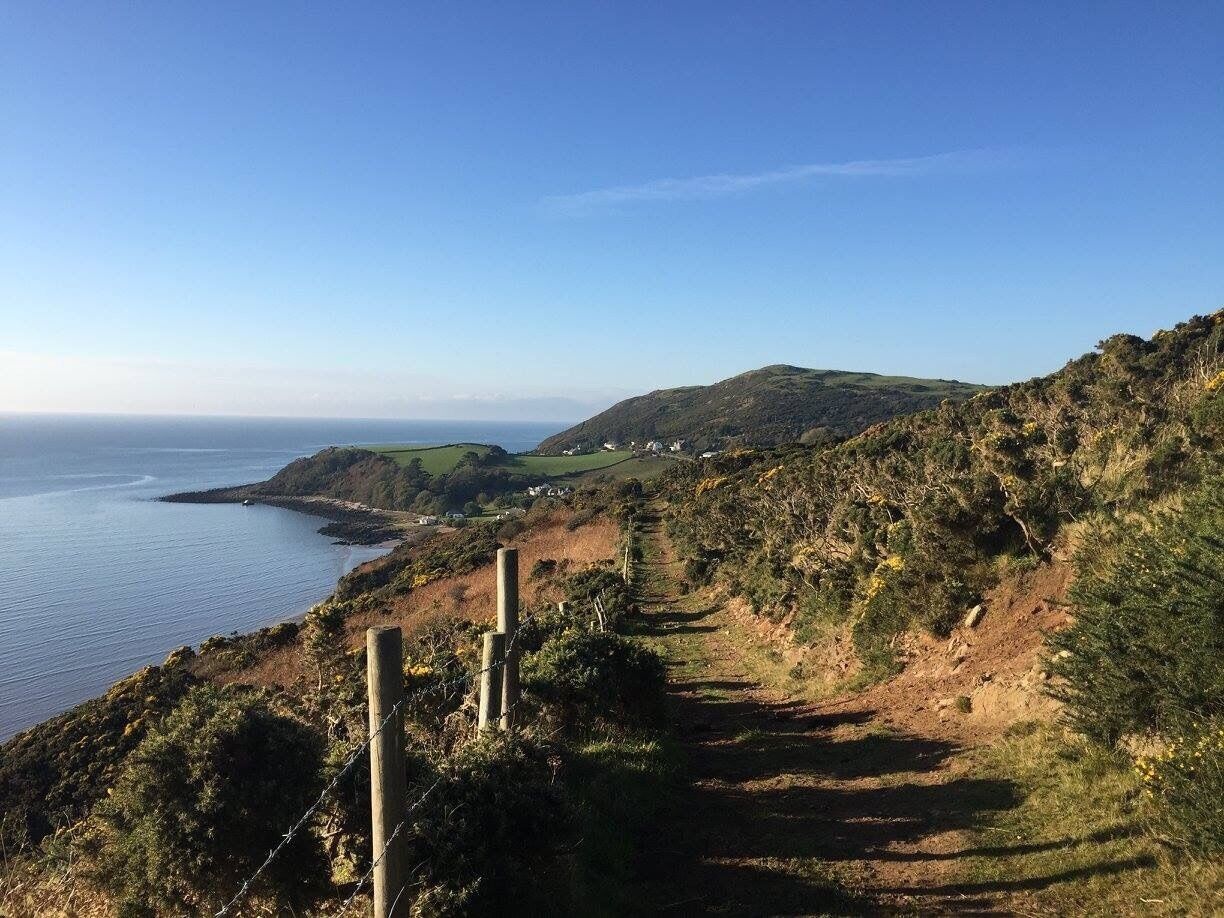 A coastal landscape with a dirt path, wooden fence posts, and a view of the sea and hills.