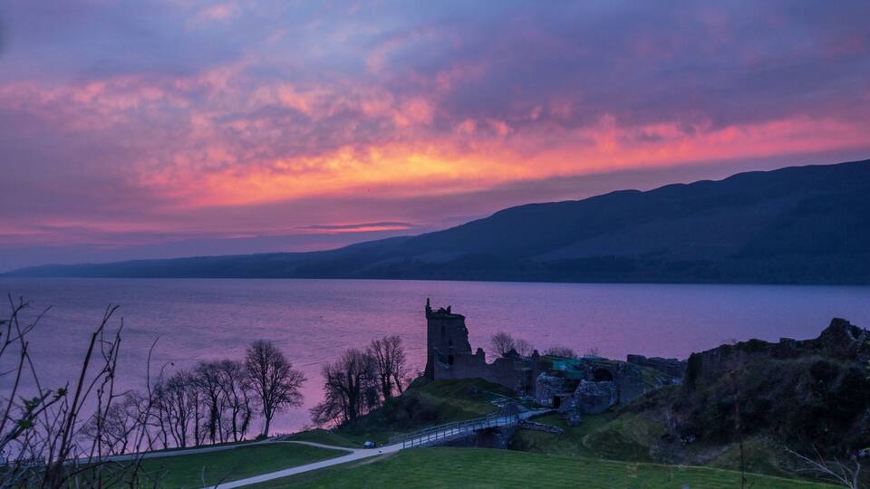 A sunset over a lake with a castle ruin and a bridge.