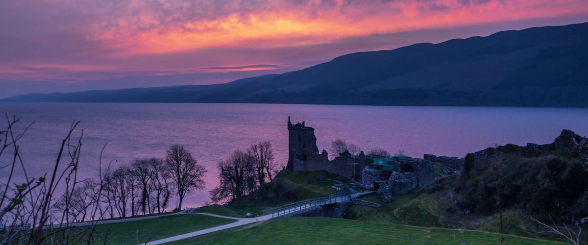 A sunset over a lake with a castle ruin and a bridge.