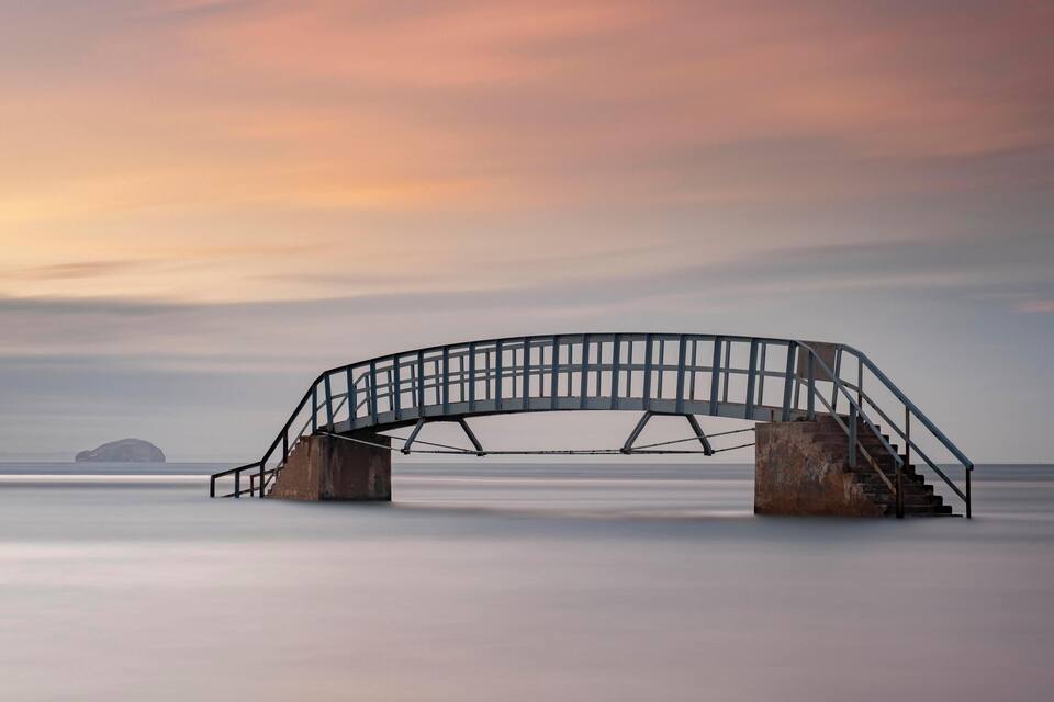 A metal bridge over calm water with a sunset sky.