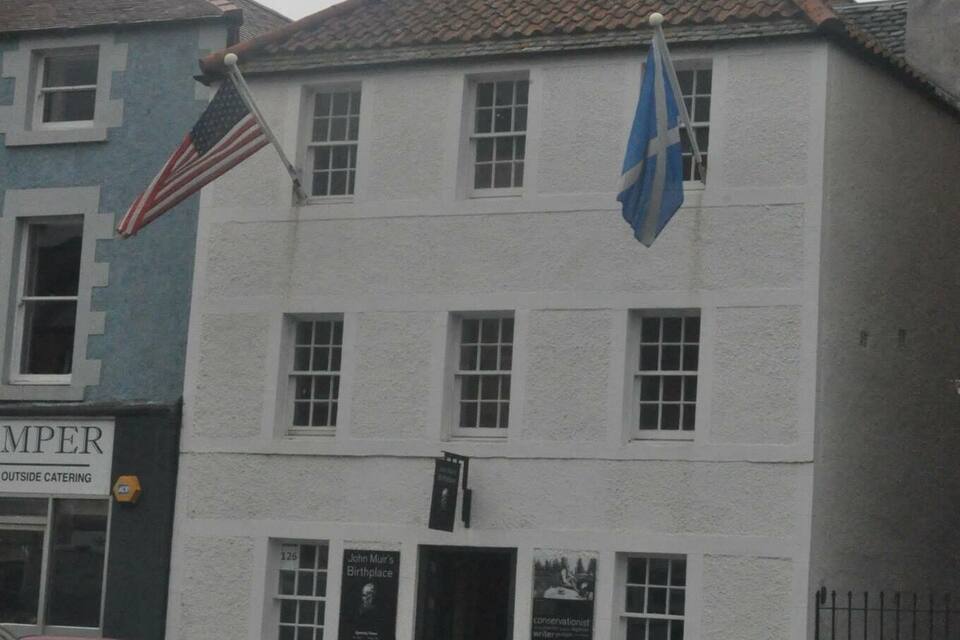 A building with a white facade and a blue flag. There are cars parked in front, and an American flag is also visible.