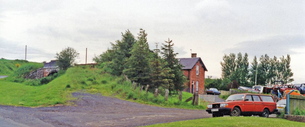 A rural road with a red car parked on the side, a brick house, and a stone bridge in the background.