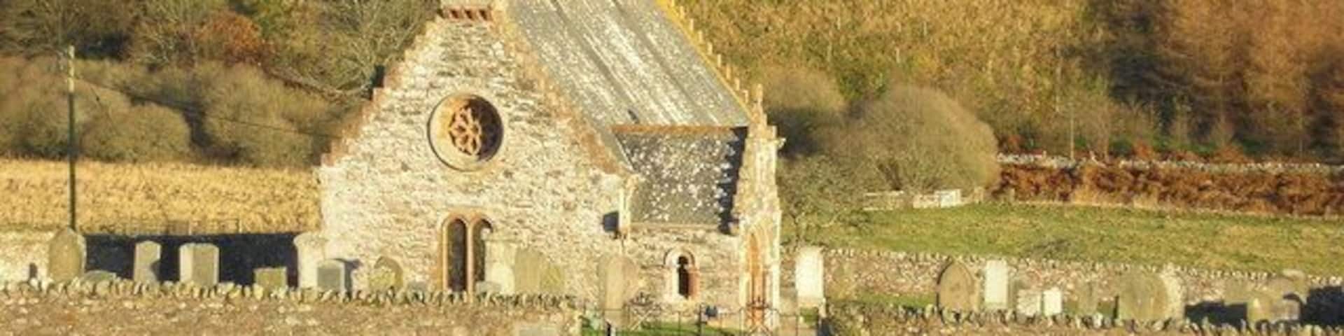 A stone church with a bell tower and a circular window, surrounded by a cemetery and a forested hill.