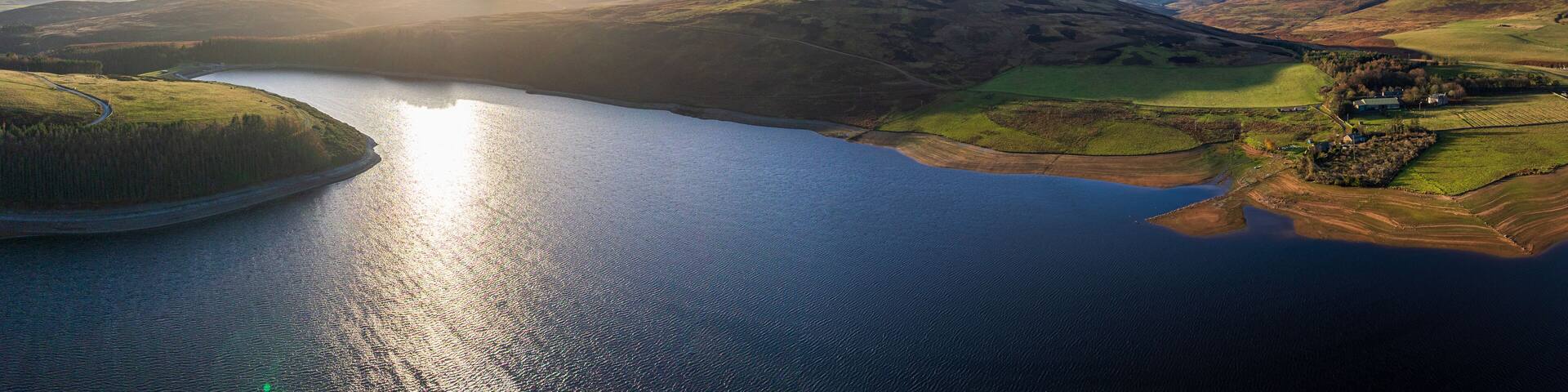 Aerial view of a serene lake reflecting the golden sunlight, embraced by rolling hills and lush green fields, Duns, Scotland, United Kingdom.