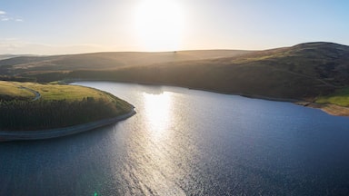 Aerial view of a serene lake reflecting the golden sunlight, embraced by rolling hills and lush green fields, Duns, Scotland, United Kingdom.