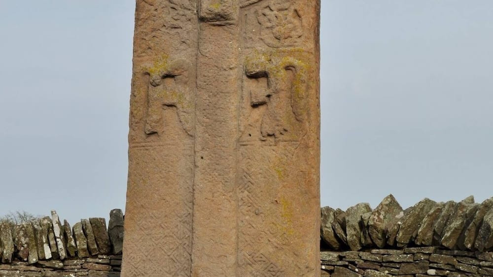 Ein hohes, uraltes Steinmonument mit geschnitzten Gesichtern und Symbolen, vor einer Steinmauer und einer grünen Wiese.