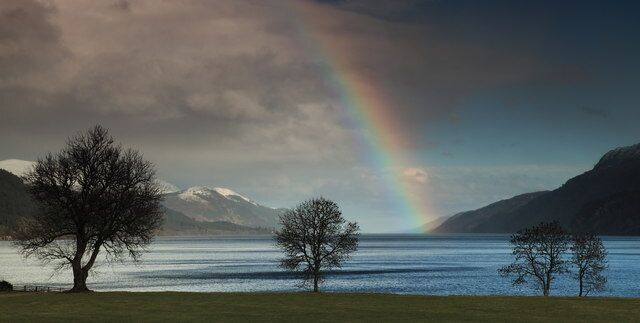 A lake with a rainbow, trees, and mountains in the background.