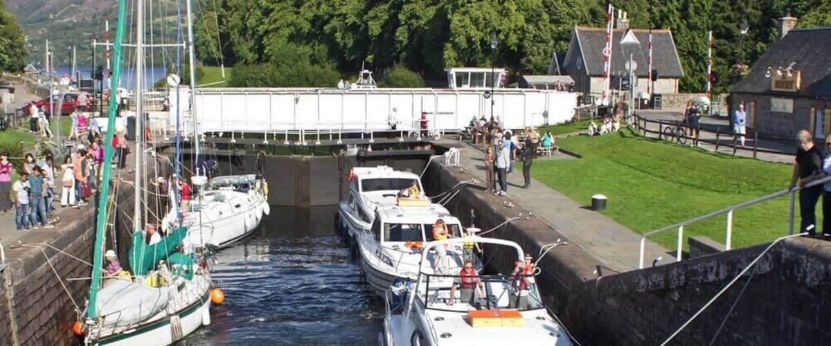 Een gracht met aangemeerde boten, een sluis en een stad met gebouwen en groen op de achtergrond.