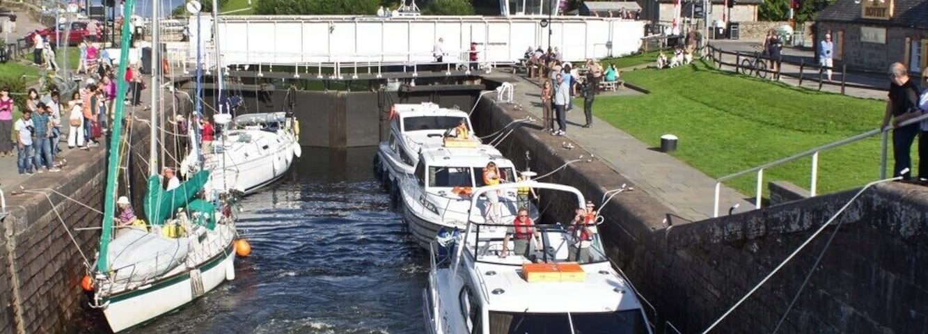 Un canal con barcos amarrados, un sistema de esclusas y un pueblo con edificios y vegetación al fondo.