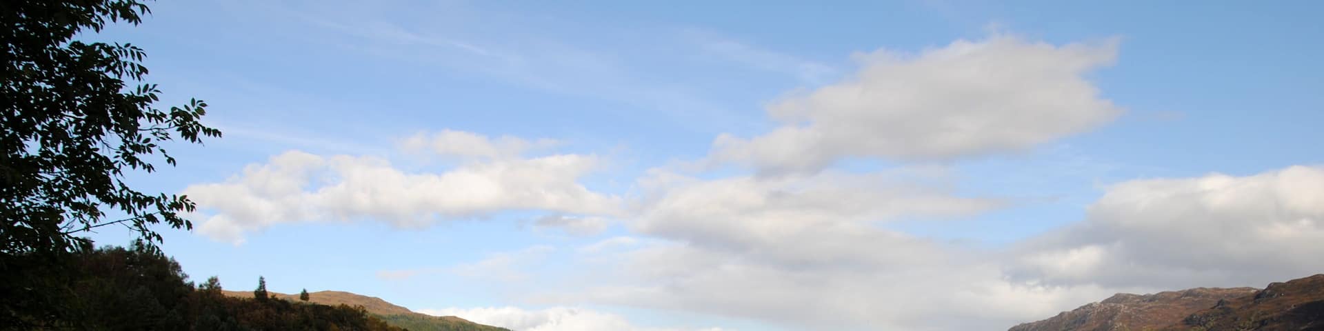A sign reading "Loch Ness Welcome to Fort Augustus" by a calm lake with hills in the background.