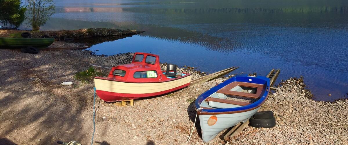 Two small boats on a pebbly shore by a calm lake, surrounded by forested hills.