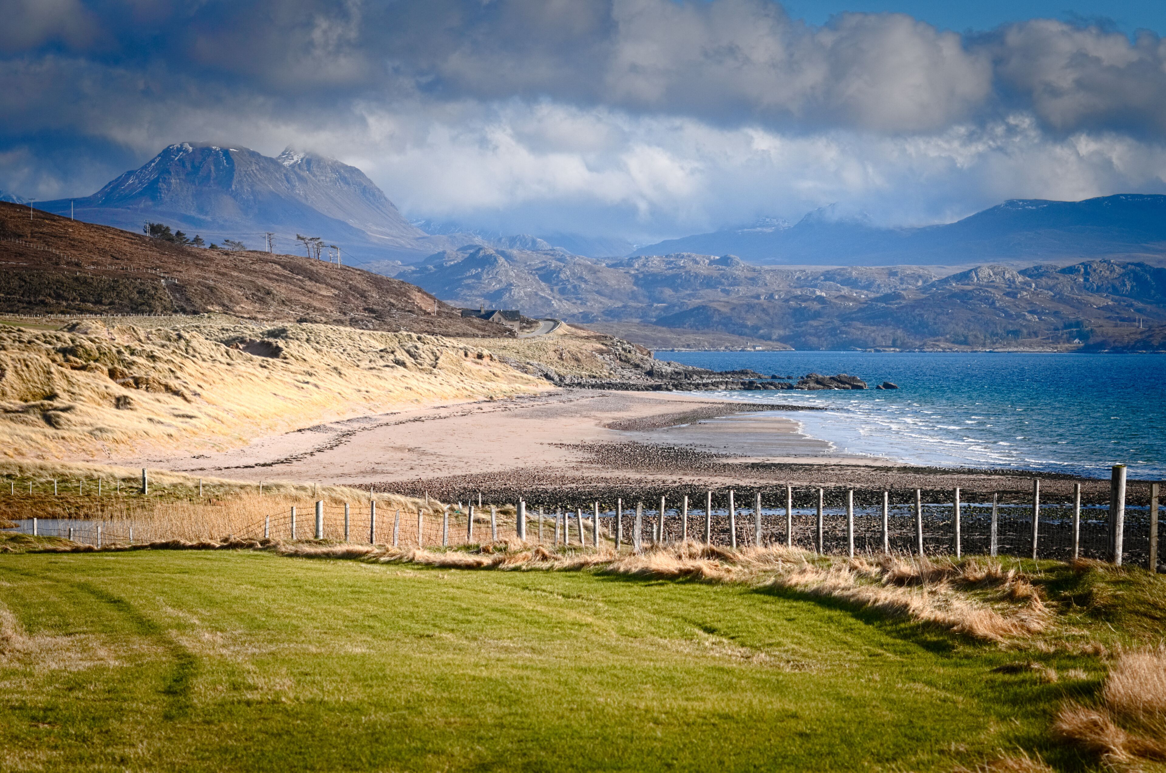 A coastal landscape with a beach, grassy fields, and mountains in the background.