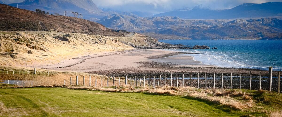 A coastal landscape with a beach, grassy fields, and mountains in the background.