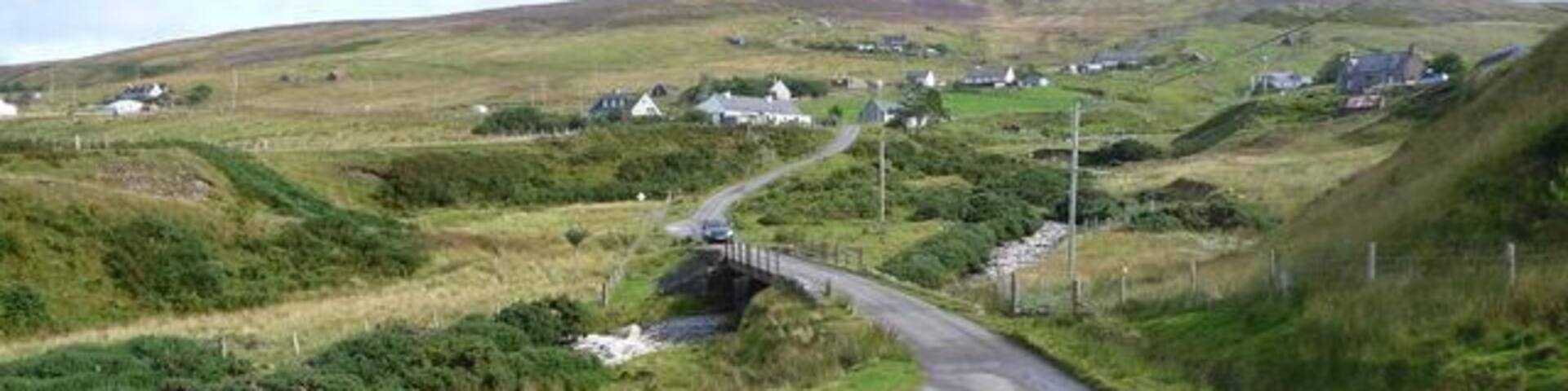 A rural road curving through a hilly landscape with scattered houses and a bridge over a stream.