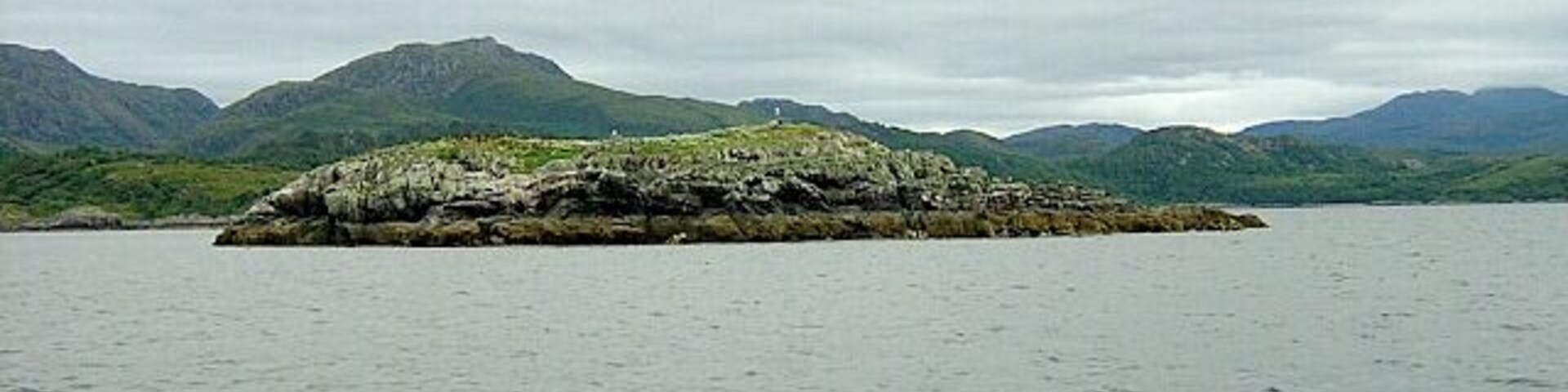 A rocky island surrounded by calm water and mountains in the background.