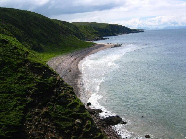 A coastal landscape with a sandy beach, green hills, and a calm sea.