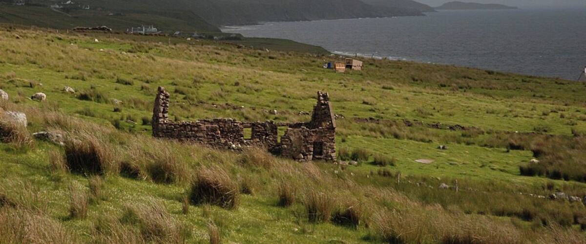 A coastal landscape with a ruined stone structure, grassy fields, and distant hills.