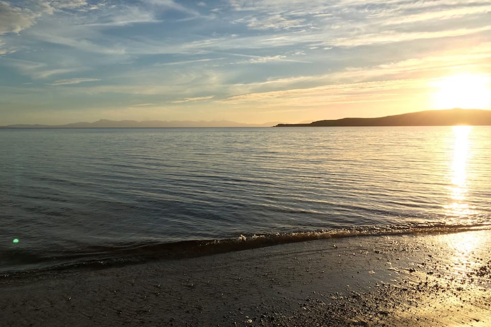 A beach with gentle waves, a clear sky, and the sun setting on the horizon.