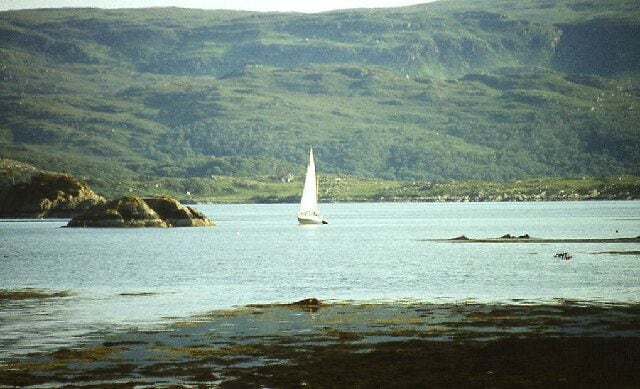 A sailboat on a calm lake with rocky outcrops and green hills in the background.