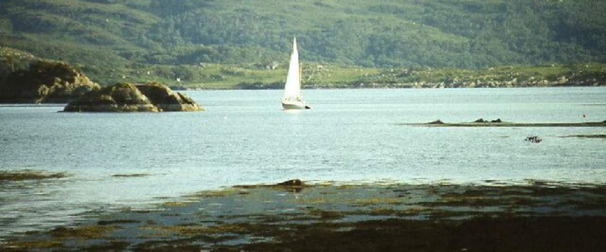 A sailboat on a calm lake with rocky outcrops and green hills in the background.