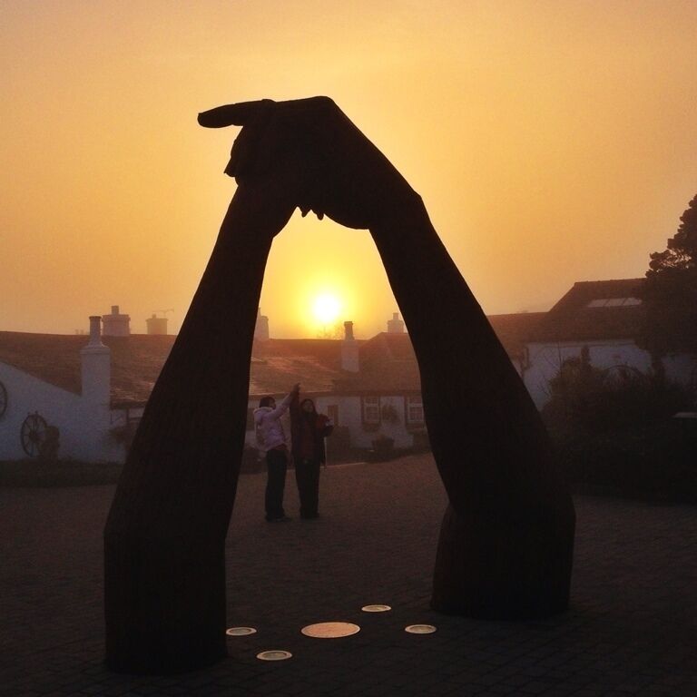 A sunset silhouette of a hand sculpture against a sky, with buildings and people in the background.