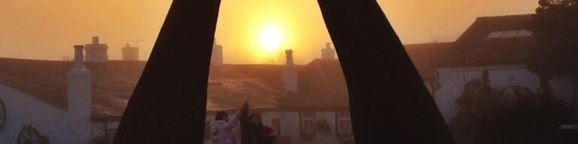 Silueta al atardecer de una escultura con forma de mano contra un cielo, con edificios y gente de fondo.
