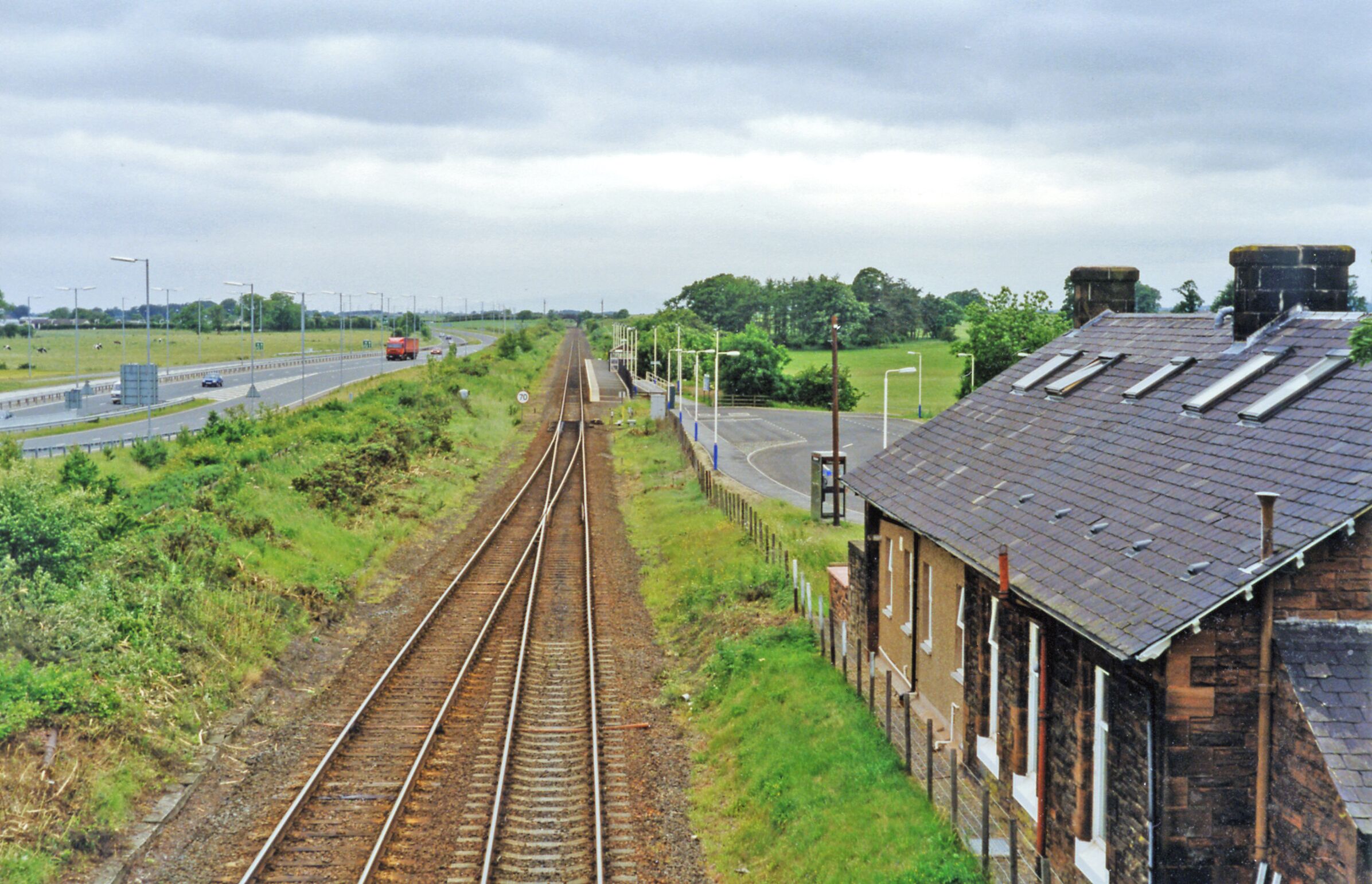 Gretna Green stations (closed and open), 1997. View westward, towards Dumfries etc.: ex-G&SWR Carlisle - Dumfries - Glasgow main line. The original station, in the immediate foreground, was closed 6/12/65 and soon after the main line was singled to Annan. However, on 20/9/93 a new station was built a short distance to the west - with a single platform as seen, but when the line was doubled again in 2008 another platform was provided on the Down side. Over to the left is the A75 road to Dumfries and Stranraer.