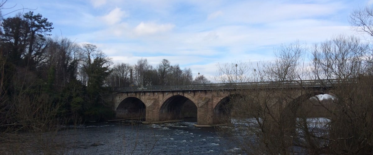 A stone bridge over a river with trees and a cloudy sky.