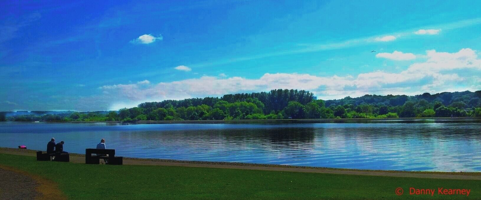 Um lago com uma margem gramada, duas pessoas sentadas em bancos e uma colina arborizada ao fundo.