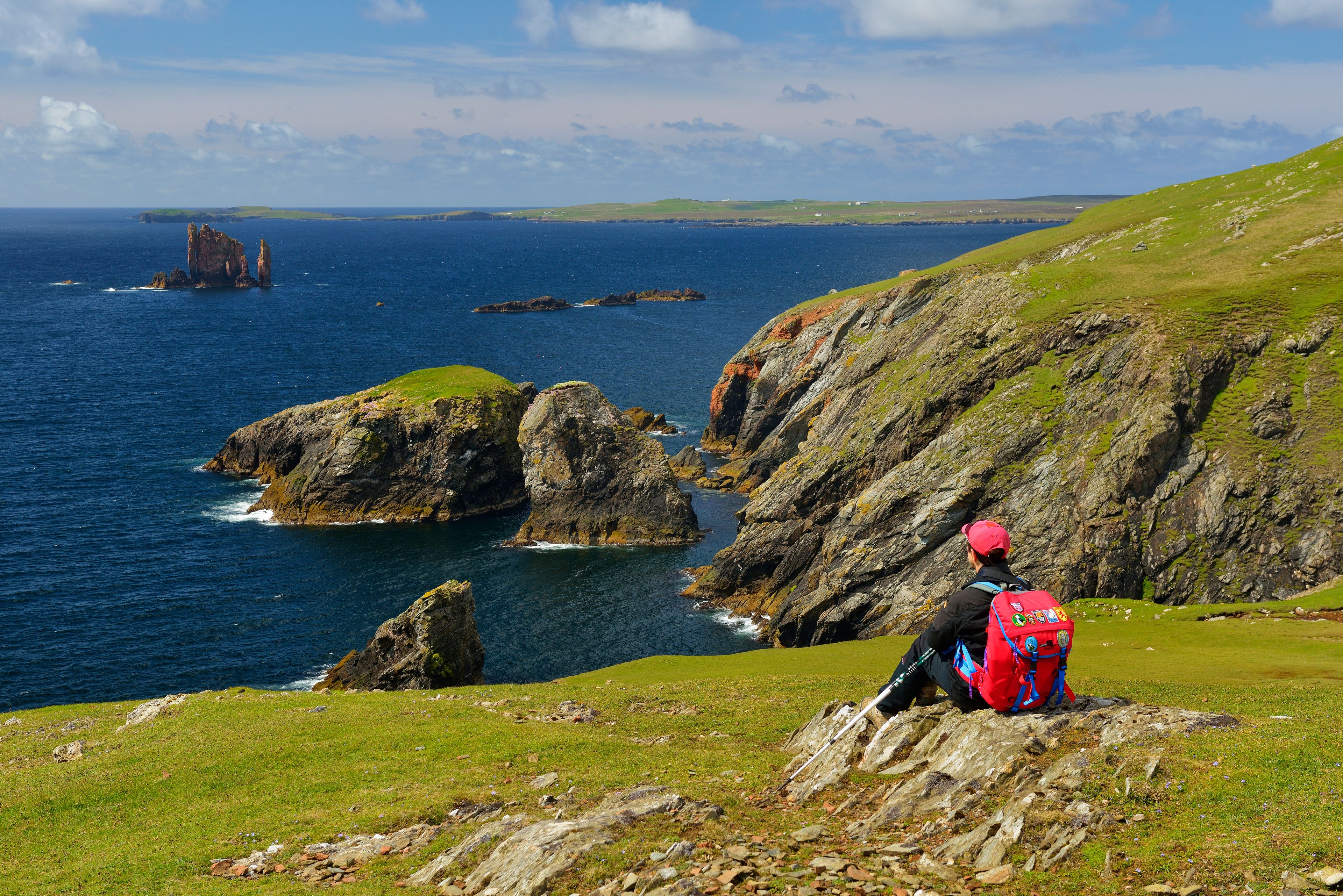 Un randonneur, portant un sac à dos rouge, se tient sur une falaise herbeuse qui surplombe une côte accidentée, parsemée de rochers et bordée par la mer.
