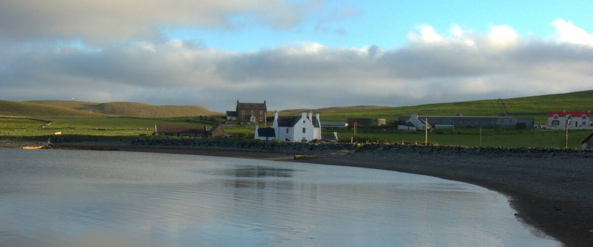 A coastal landscape with a sandy beach, calm water, and a white house with a red roof in the background.
