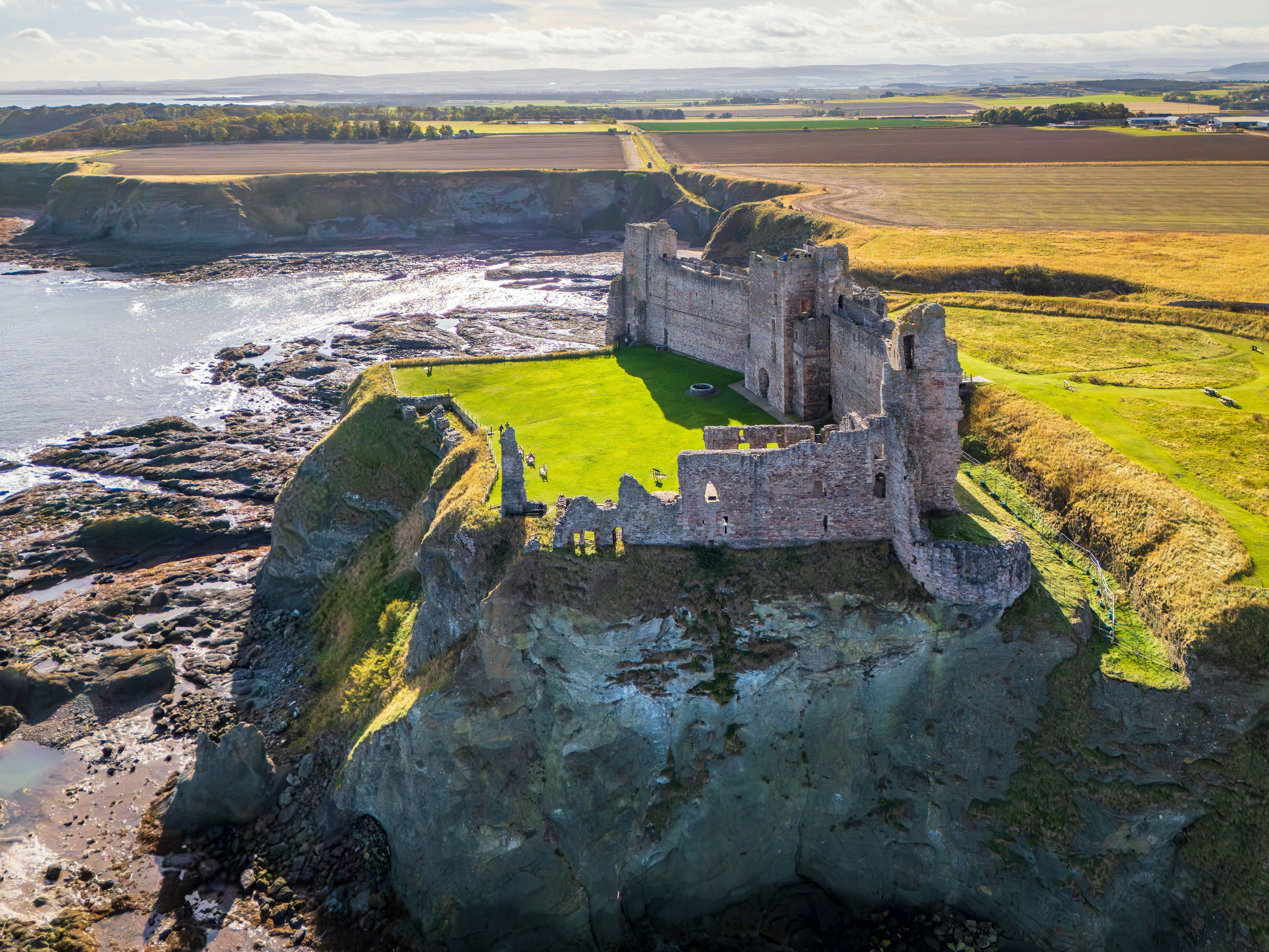 Un château perché sur une falaise, surplombant une zone côtière avec des champs en arrière-plan.