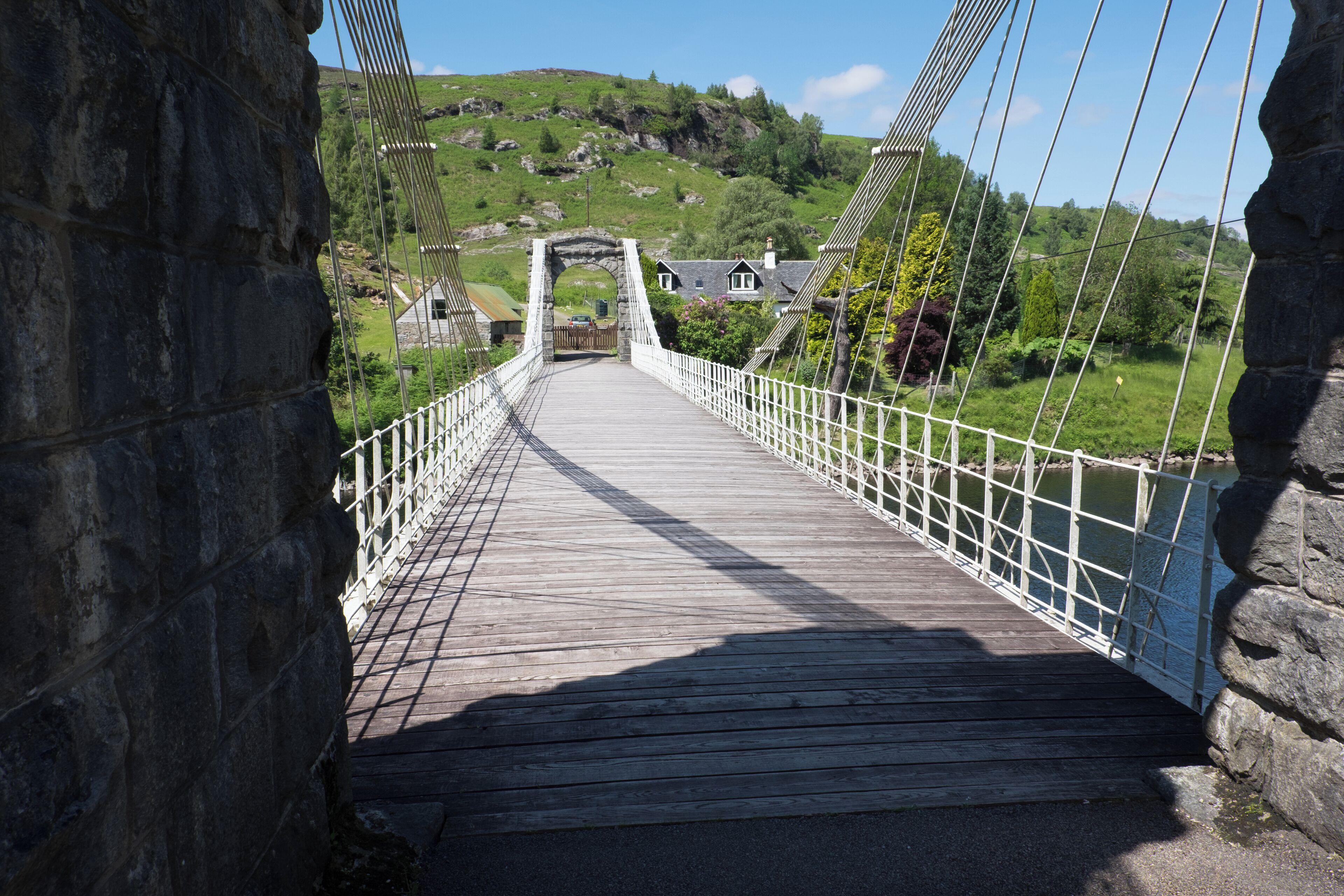 A wooden suspension bridge with white railings, leading to a stone archway and houses on a hillside.