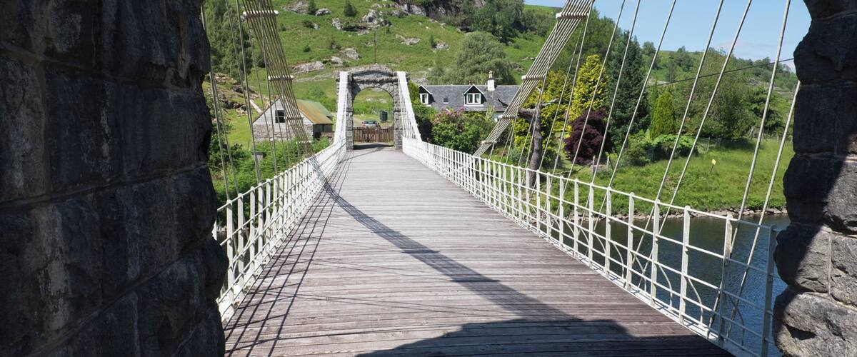 A wooden suspension bridge with white railings, leading to a stone archway and houses on a hillside.