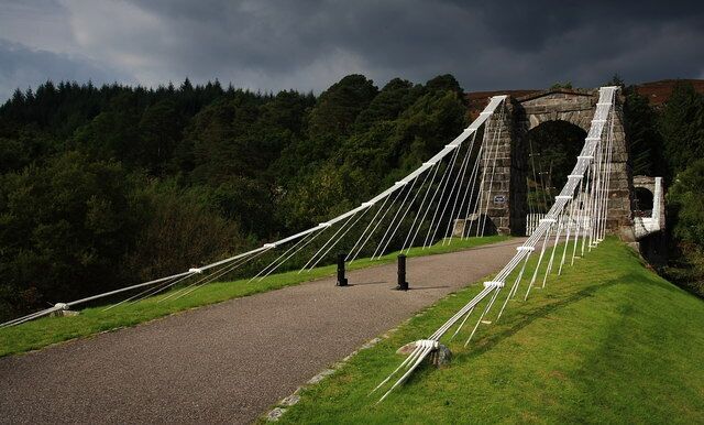 A suspension bridge with cables and a stone arch bridge in the background.