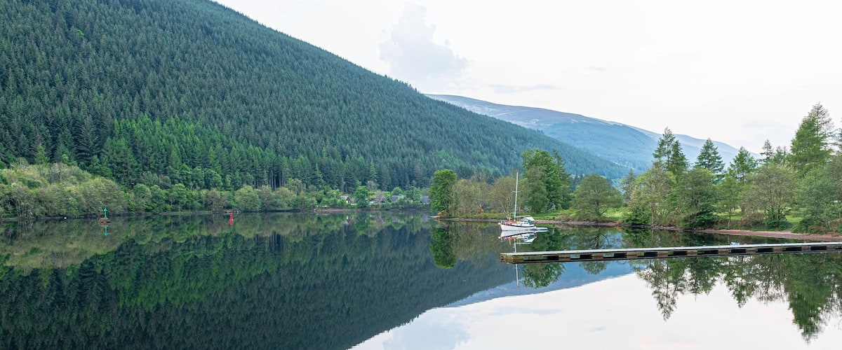 A calm lake with a boat docked on the right, surrounded by dense forest and mountains in the background.