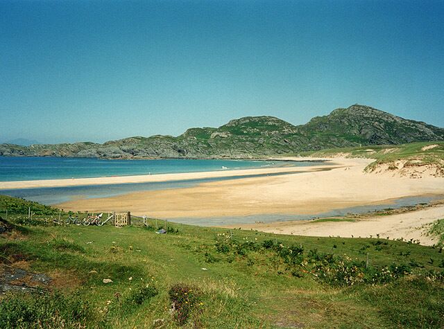 A coastal landscape with a sandy beach, grassy foreground, and distant hills.
