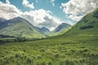 A valley with lush green fields, rolling hills, and mountains under a partly cloudy sky.