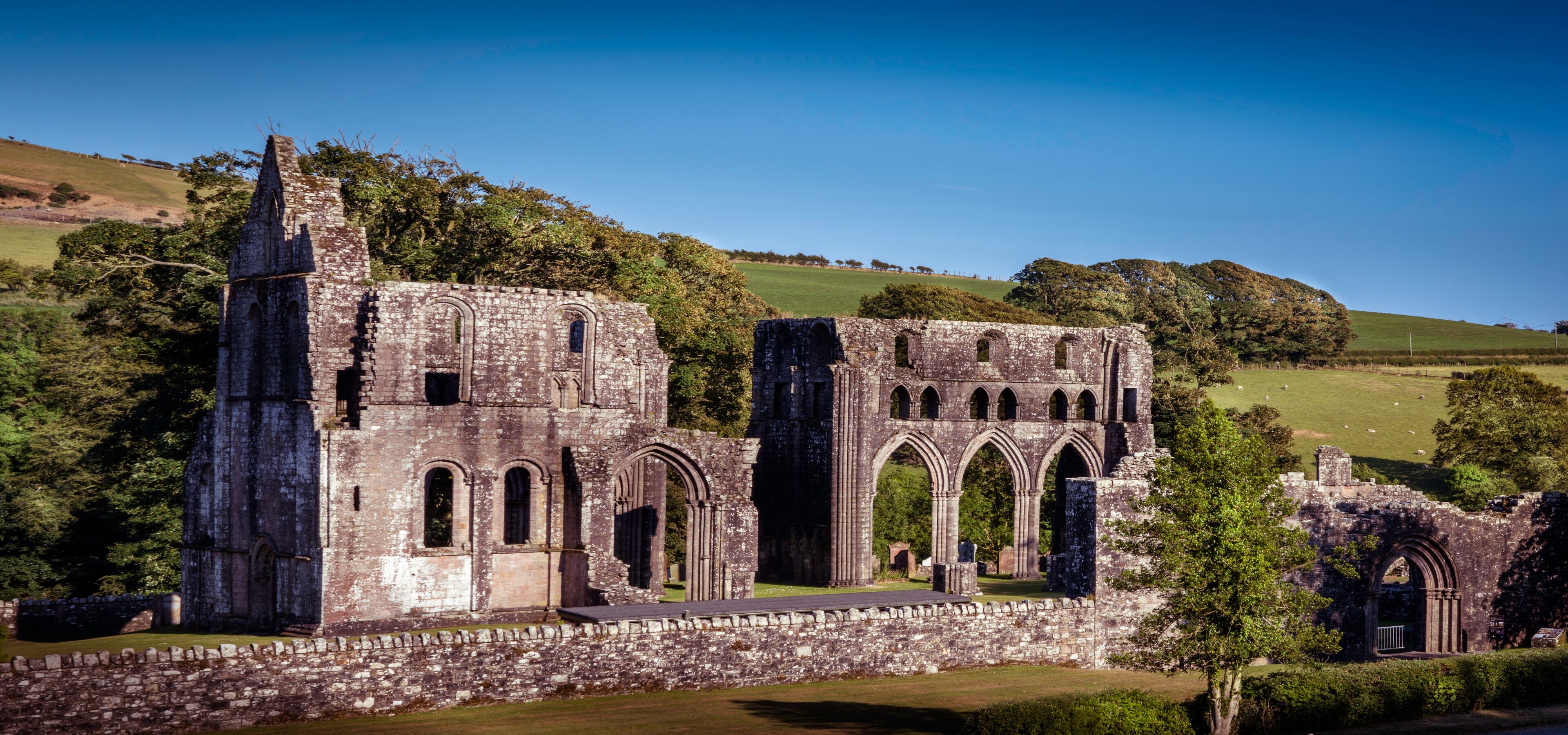 An ancient stone structure with arched windows and a tower, surrounded by lush green fields and trees.