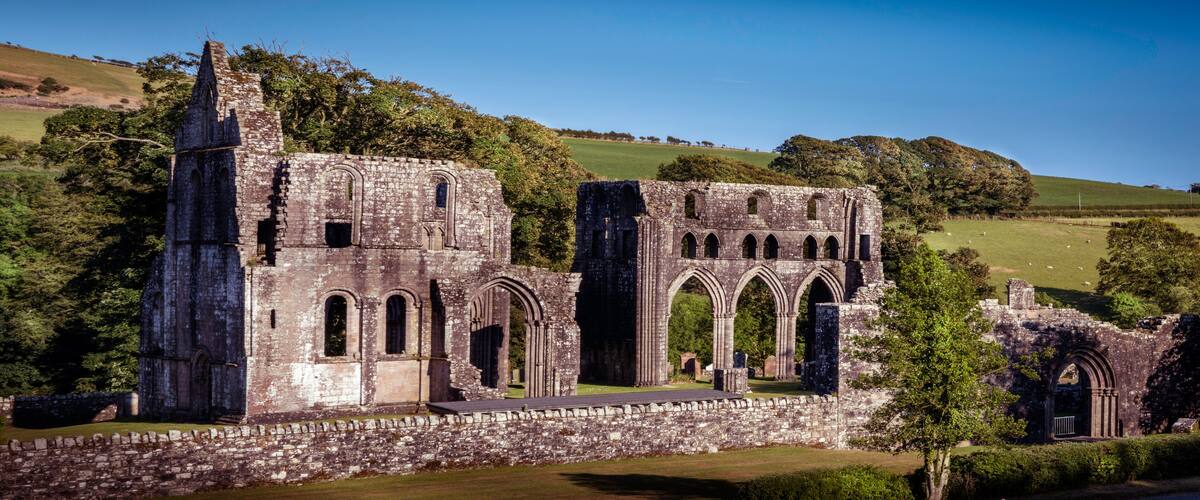 An ancient stone structure with arched windows and a tower, surrounded by lush green fields and trees.