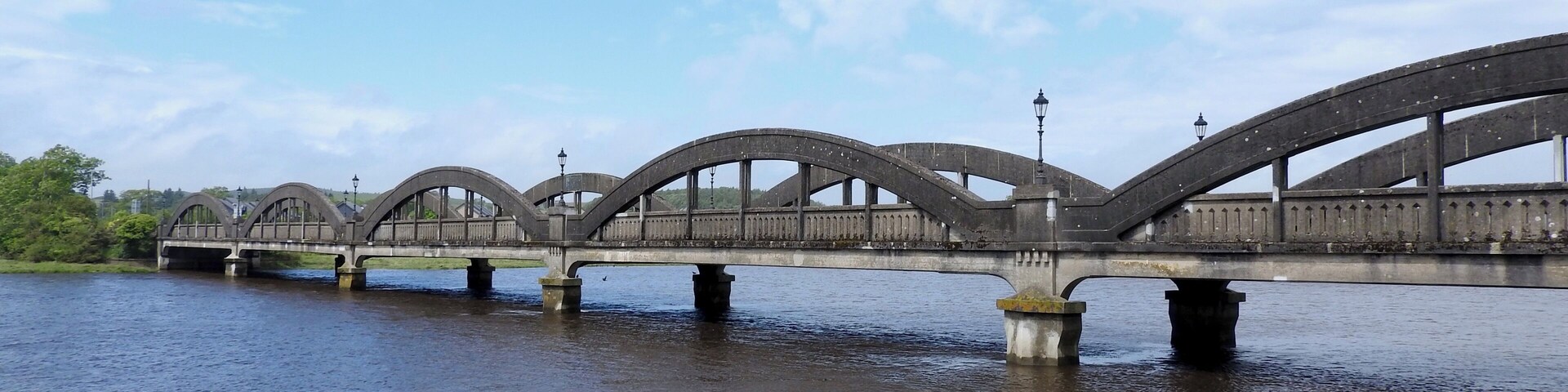 The bridge over the River Dee at Kirkcudbright was built in 1926 by the engineers Mouchel. It provided a direct route west out of the town, rather than having to head north via Tongland Bridge first. However, old maps suggest that there may have been an earlier bridge on the site.
The bridge is a massive 5 span reinforced concrete structure, where each span is supported by tied arch trusses overhead. Unlike many other similar structures, the arches are not tied across the carriageway, mainly because they are too shallow to then allow traffic through! Below the spans the piers are paired and while only two stand in the river at low tide, at high tide all are partially submerged.
The bridge is signed as a Weak Bridge with a 17T weight limit, and for some unknown reason, has Wig-Wag signs at either end.
