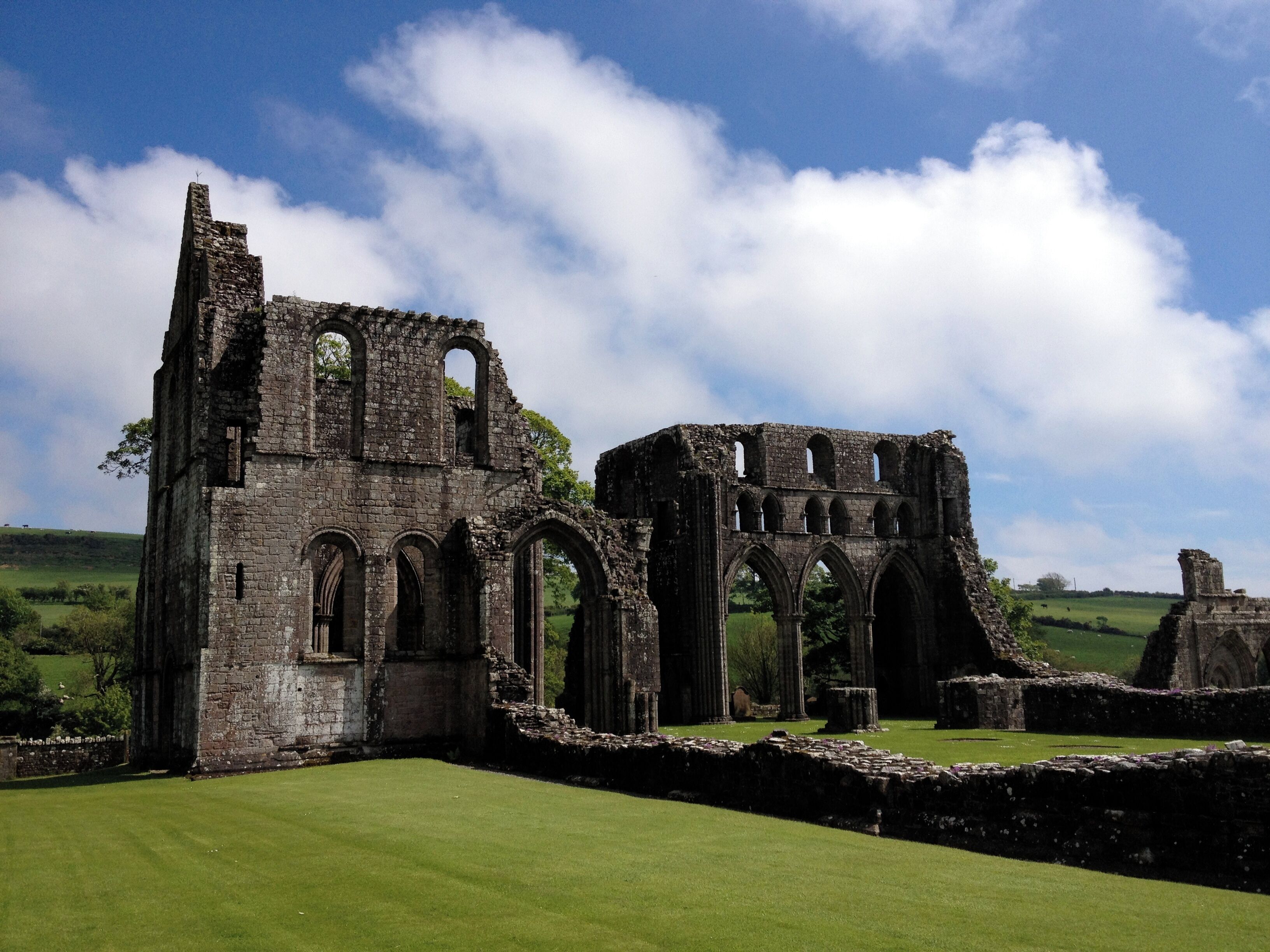The ruins of a medieval stone structure with arches and columns, surrounded by green grass and a clear sky.