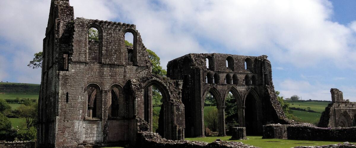 The ruins of a medieval stone structure with arches and columns, surrounded by green grass and a clear sky.