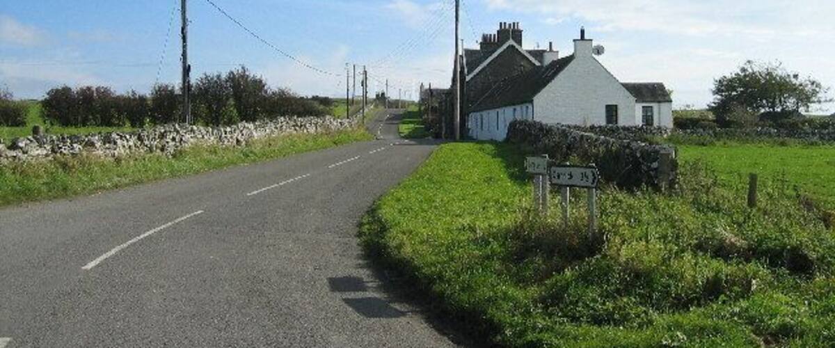 A rural road with a white house on the right, surrounded by greenery and a stone wall.