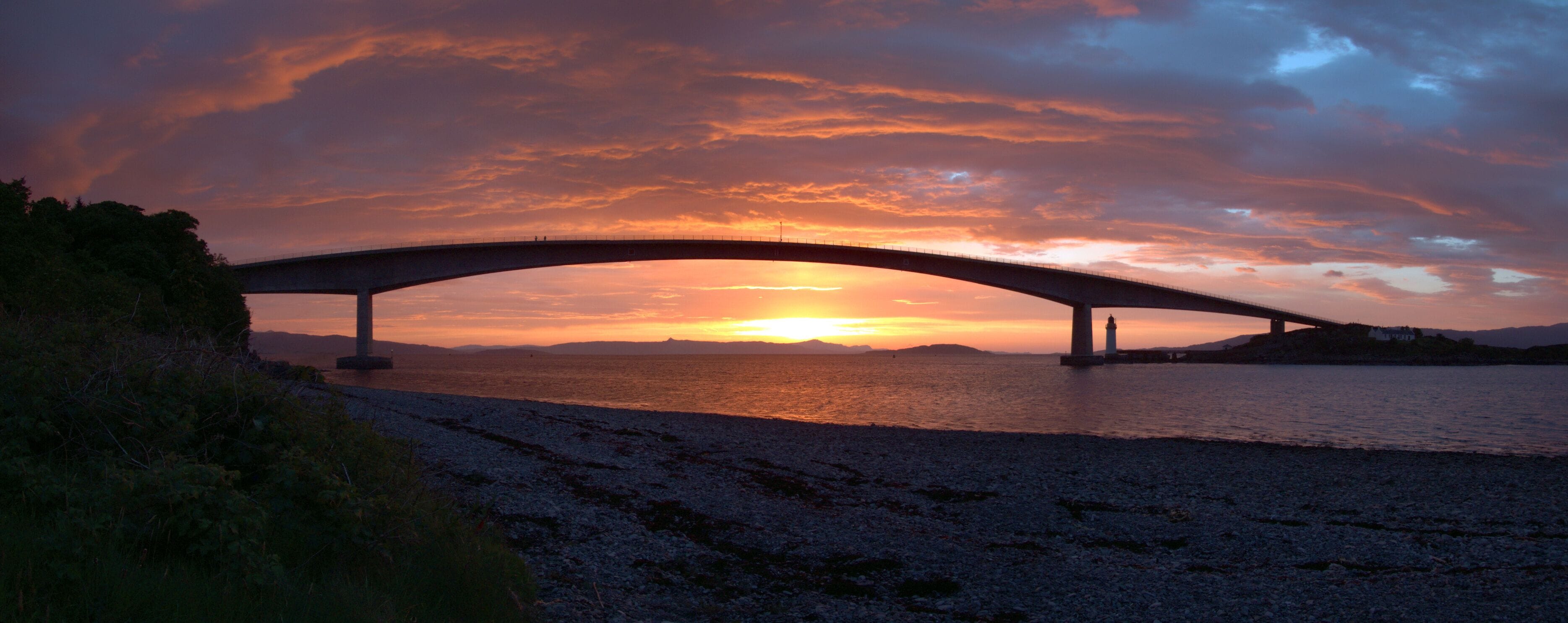 A bridge over a body of water at sunset with a lighthouse in the distance.