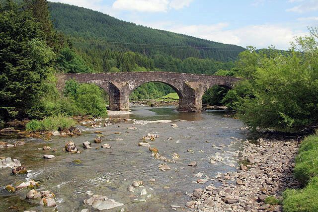 A stone bridge over a river with rocky banks and green forest in the background.