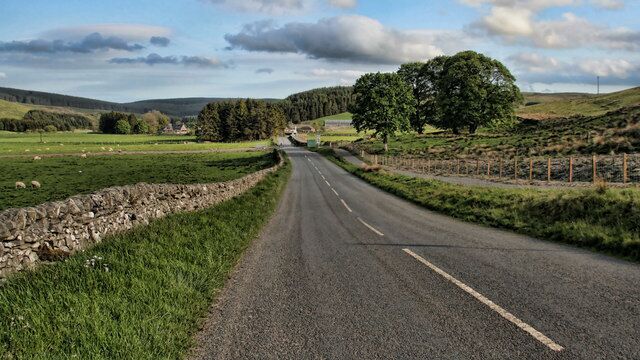 A rural road flanked by stone walls and green fields, leading through a hilly landscape with trees and a clear sky.