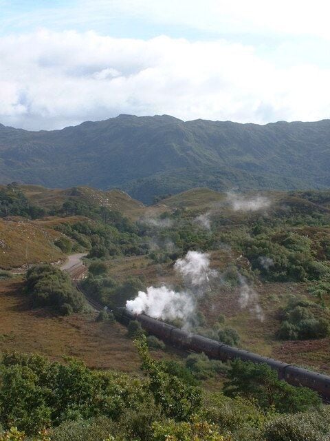 A steam train in a mountainous landscape with greenery and a clear sky.