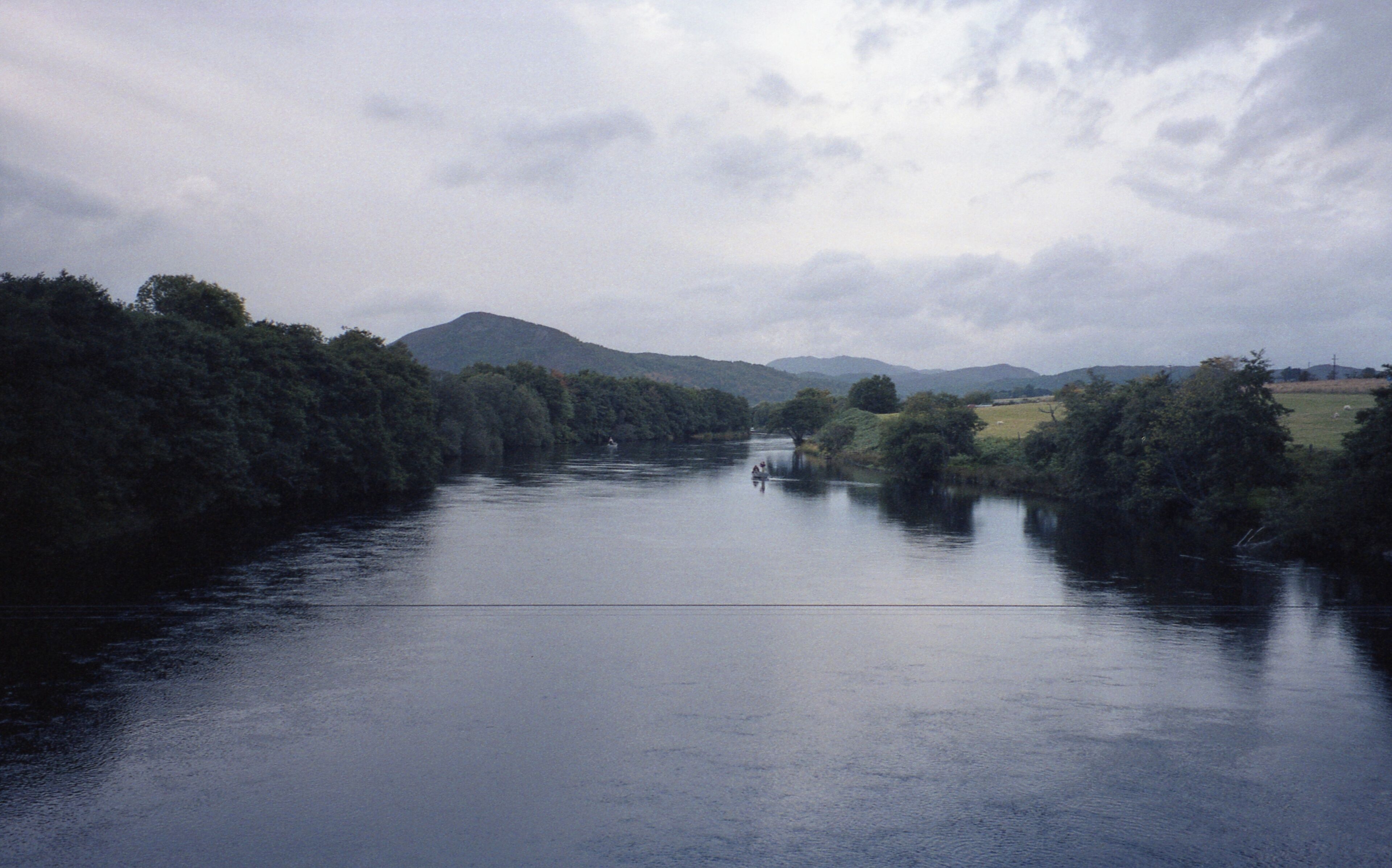 A river flanked by dense forests and distant mountains under a cloudy sky.